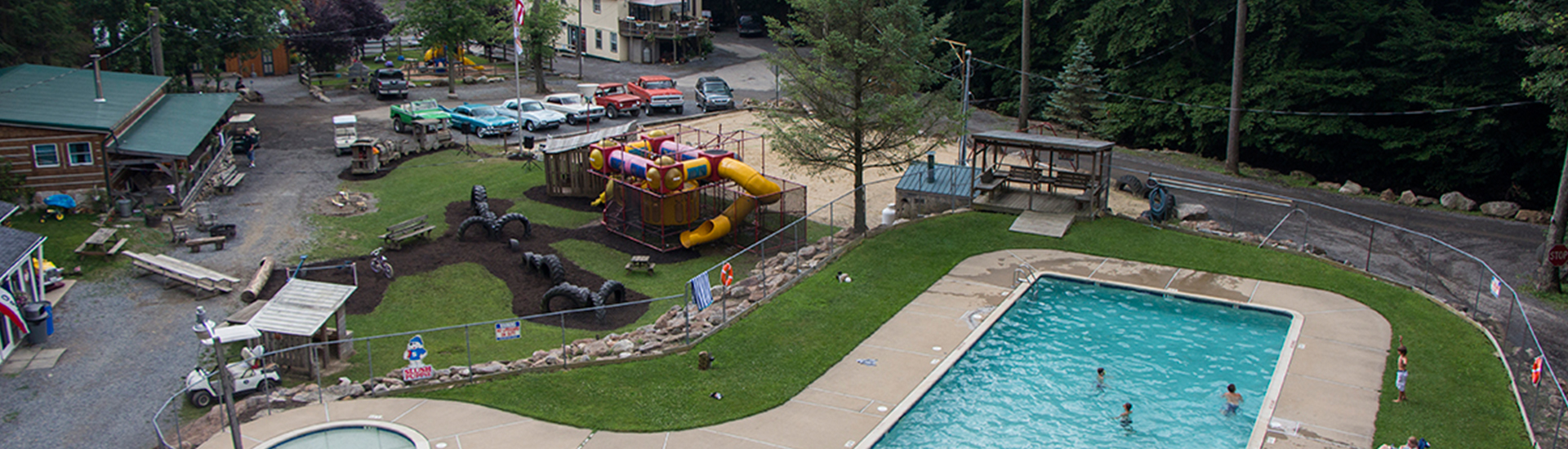 A picture of our campground. There is a line of classic cars by the office. Nearby is the playground, and a sandy area for playing volleyball. In the foreground is a swimming pool with kids swimming.