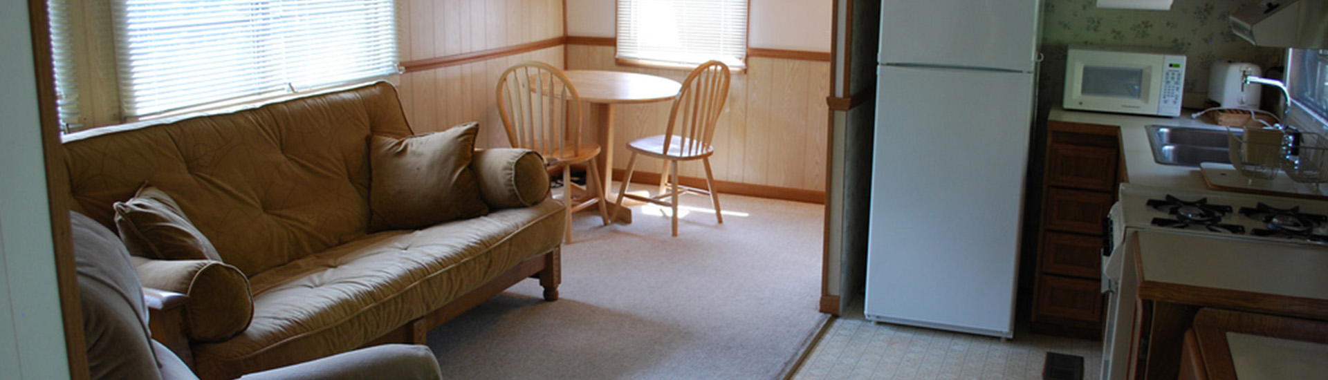 Inside of a cottage, with a sofa on the left, a table and chairs nearby, and the kitchen to the right.